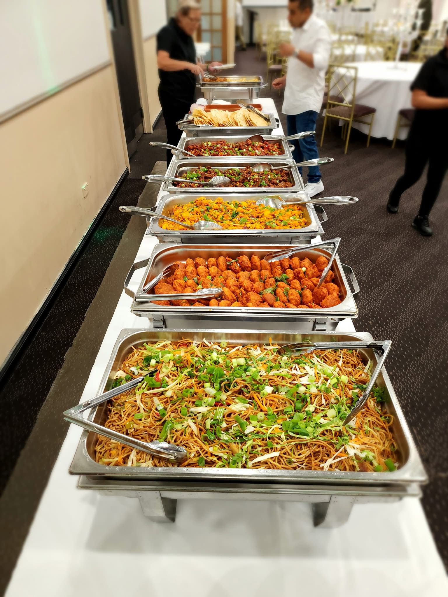 Stainless steel chafing dishes set up on a table with various Indian and Nepalese dishes. Dish items include noodles garnished with herbs, pakoras, biryani, curries, chicken, naan, and vegetable dishes. Buffet-style setup with professional catering equipment at Third Eye Windsor, 515 George St, South Windsor NSW 2756.