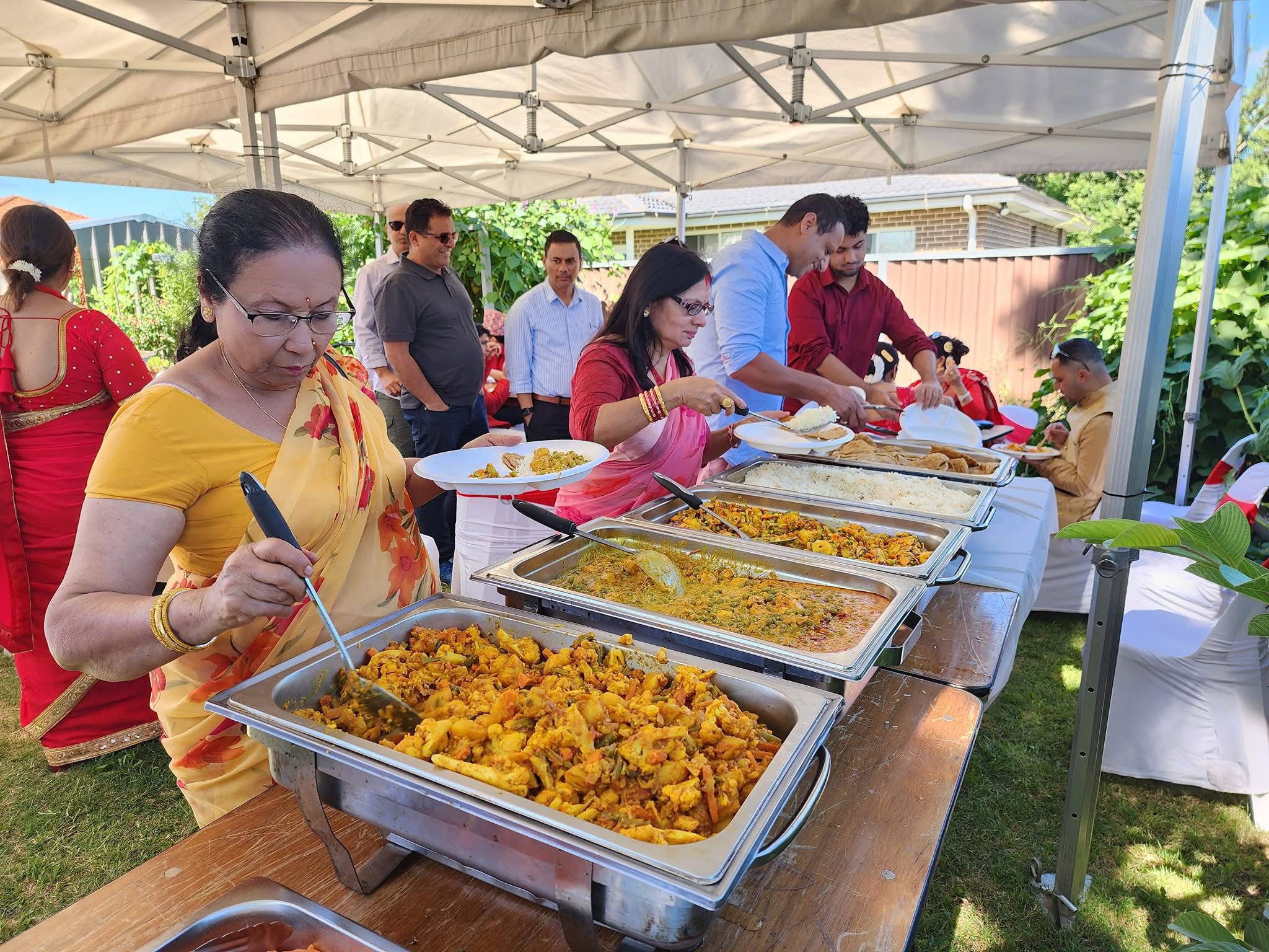 Outdoor family event of a Nepali family with catering service provided. Several people are seated under large white canopy tents on a grassy area. Attendees are enjoying meals on plates at tables covered with white tablecloths at Third Eye Windsor, 515 George St, South Windsor NSW 2756.