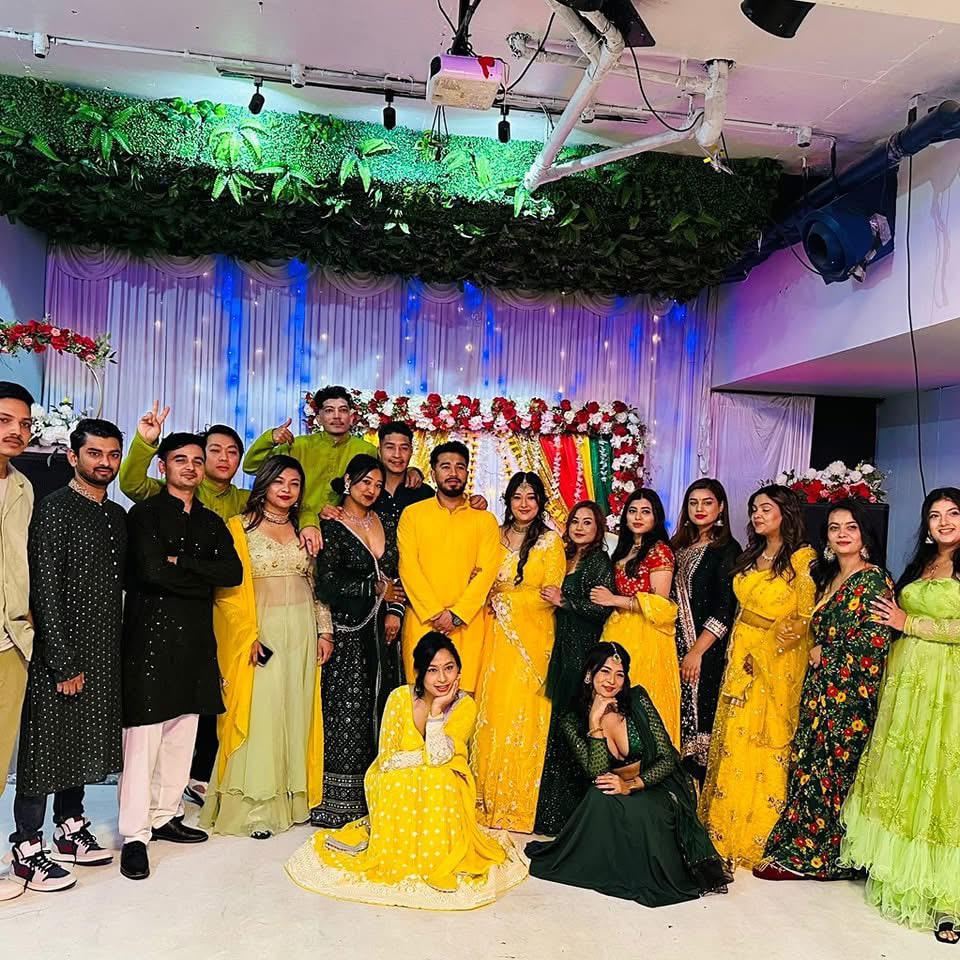 A large group of people in formal traditional attire, many wearing yellow or gold outfits, others in black or patterned clothes. They are posing in front of a decorated backdrop with white curtains, blue lighting, and floral arrangements. Indoor event space with greenery hanging from the ceiling at Third Eye Windsor, 515 George St, South Windsor NSW 2756.