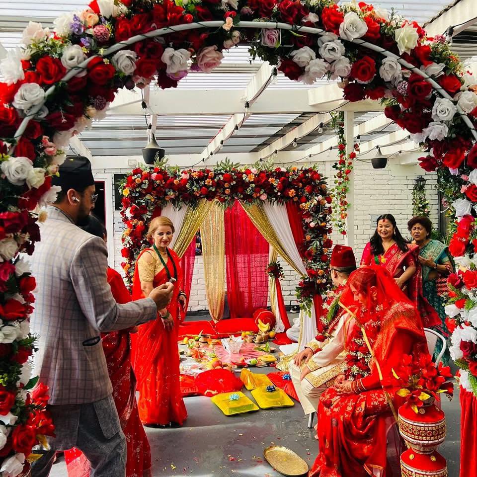 Nepalese wedding ceremony indoors at Third Eye Windsor with catering service. A decorated mandap with a red and white rose arch. Traditional floor seating with red cushions and yellow plates for rituals. Ceremonial items arranged on the floor. People in traditional clothes. Bride in red dress with gold details. Groom in traditional daura suruwal at Third Eye Windsor, 515 George St, South Windsor NSW 2756.