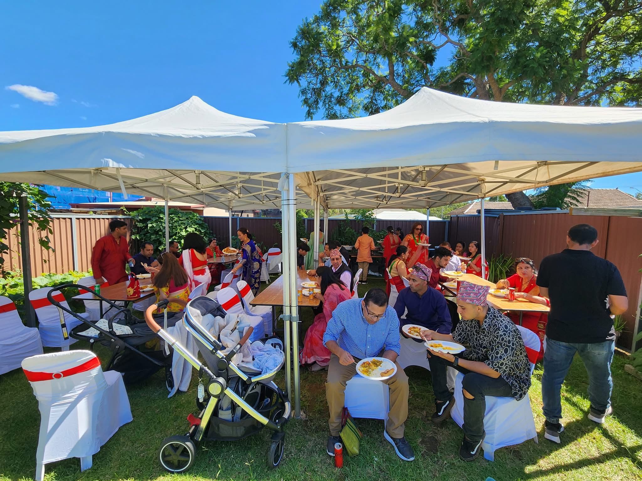 Outdoor family event of a Nepali family with catering service provided. Several people are seated under large white canopy tents on a grassy area. Attendees are enjoying meals on plates at tables covered with white tablecloths at Third Eye Windsor, 515 George St, South Windsor NSW 2756.